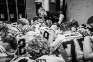 Football team huddles with coach in locker room.