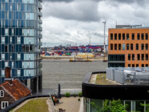 Modern buildings overlook a busy port with cranes and ships.