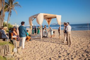 Beach wedding ceremony with guests under a canopy.