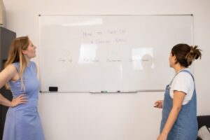 two women standing in front of a white board