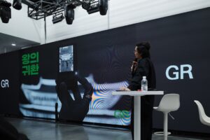A person speaks at a presentation with a large screen.