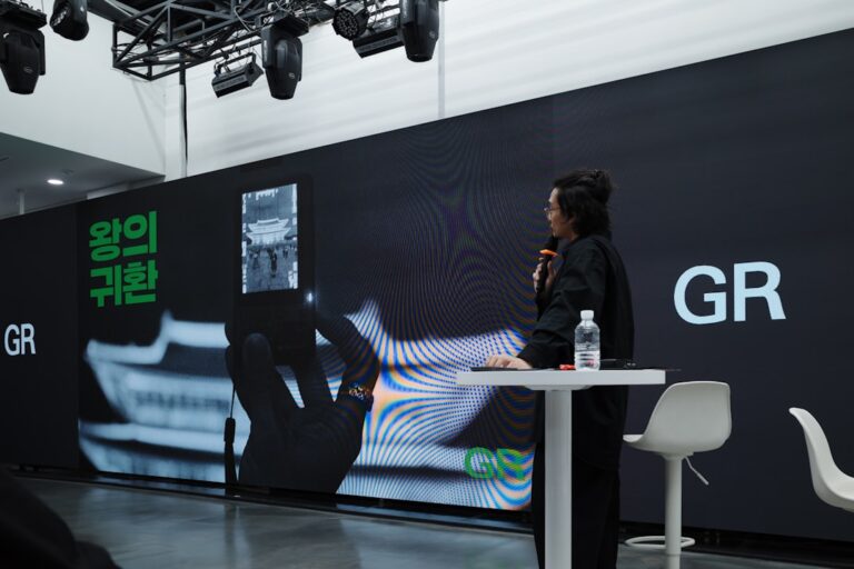 A person speaks at a presentation with a large screen.