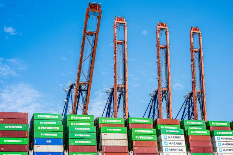 Port cranes and stacked shipping containers under blue sky.