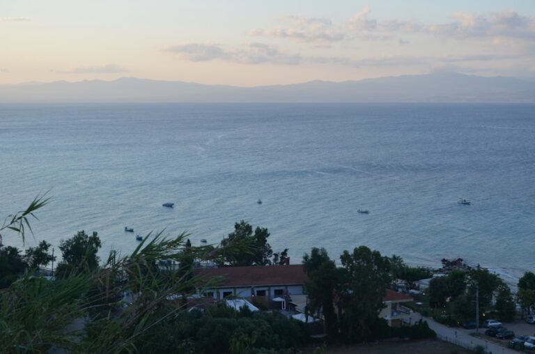 Coastal view with calm sea, buildings, trees, and distant mountains.