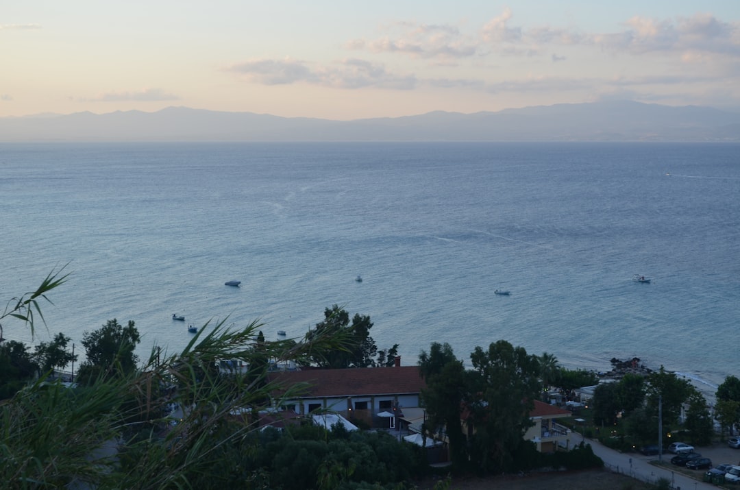 Coastal view with calm sea, buildings, trees, and distant mountains.